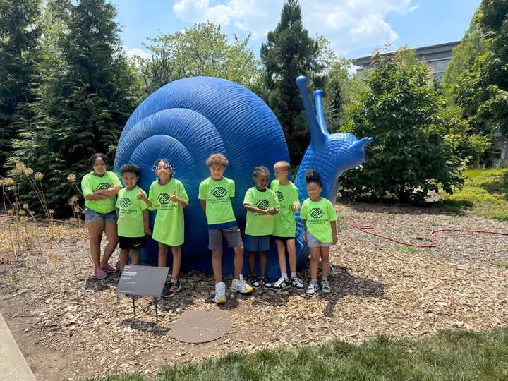 Club members in green Boys and Girls Clubs shirts posing beside a large blue inflatable snail sculpture on a summer outing