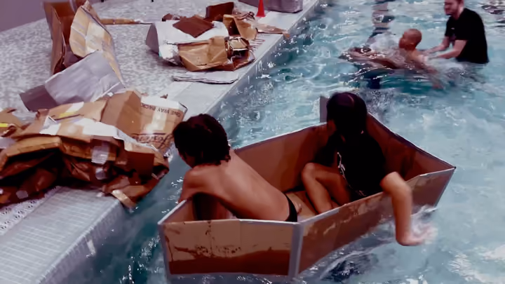 Two campers paddling a cardboard boat across an indoor pool, with soaked cardboard piled on the deck behind them